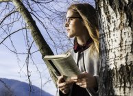 Молодая женщина стоит рядом с деревом, держа книгу — стоковое фото