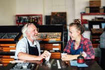 Senior-Handwerker plaudert und lacht mit junger Kunsthandwerkerin in Buchkunstwerkstatt — Stockfoto