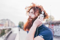 Jovem hipster masculino com cabelo vermelho e barba ouvindo fones de ouvido com os olhos fechados na cidade — Fotografia de Stock