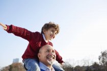 Grandson sitting on grandfathers shoulders, arms out smiling — Stock Photo
