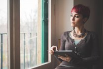 Молода жінка сидить біля вікна, використовуючи цифровий планшет — стокове фото