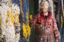 Männlicher Schiffsmaler liest Smartphone-Text, der sich gegen eine mit Farbe bespritzte Wand lehnt — Stockfoto