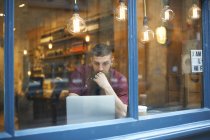 Window view of young man using laptop in cafe — Stock Photo