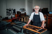 Ritratto di artigiano anziano accanto a vassoio di lettere tipografiche in officina di stampa — Foto stock