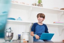 Niño sentado en la superficie de trabajo de la cocina, utilizando el ordenador portátil - foto de stock