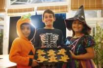 Brothers and sister wearing halloween costumes holding tray of gingerbread men — Stock Photo