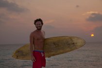 Surfista carregando bordo na praia ao pôr do sol — Fotografia de Stock