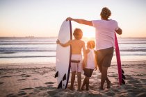 Батько і два сини, що стоять на пляжі, з дошками для серфінгу, дивляться на океан, вид ззаду — стокове фото