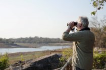 Homem sênior olhando através de binóculos no rio, Parque Nacional Kafue, Zâmbia — Fotografia de Stock