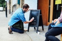Junger Mann kniet auf Gehweg und wischt Tafel vor Werkstatt — Stockfoto