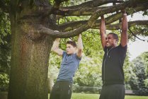 Entrenador personal y joven haciendo pull ups usando la rama del árbol del parque - foto de stock