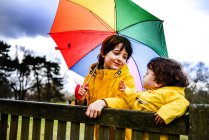 Vista aérea del bebé y el hermano en anoraks amarillos con paraguas en el banco del parque - foto de stock