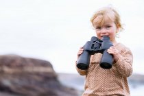 Toddler girl holding binoculars and looking at camera on beach — Stock Photo
