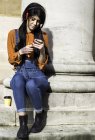 Jovem mulher sentada ao lado do pilar, usando fones de ouvido, segurando smartphone — Fotografia de Stock
