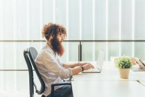 Young male hipster with red hair and beard typing on laptop at desk — Stock Photo
