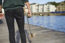 Young man walking beside river, holding skateboard, rear view, mid section, Bristol, UK — Stock Photo