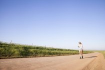Mujer adulta caminando por la carretera del campo, mirando el teléfono móvil - foto de stock
