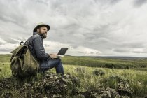 Senderista masculino maduro sentado en las rocas con ordenador portátil con vistas al paisaje, Cody, Wyoming, EE.UU. - foto de stock