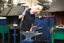 Young female trainee blacksmith hammering horseshoe on workshop anvil — Stock Photo
