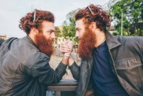 Young male hipster twins with red hair and beards arm wrestling on bridge — Stock Photo