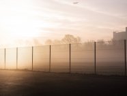Tennis court wire fence and overhead airplane in misty park at sunrise — Stock Photo