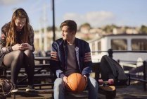 Два друга, сидящие снаружи, молодой человек, держащий баскетбол, молодая женщина, использующая смартфон, Бристоль, Великобритания — стоковое фото