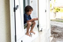 Garçon assis sur le rebord de la fenêtre de l'appartement de vacances regardant bracelet, France — Photo de stock