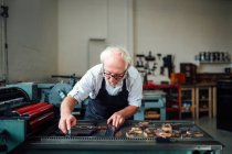Artesanato sênior curvando-se e usando letras de madeira e máquina de tipografia em oficina de artes do livro — Fotografia de Stock