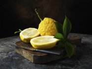 Textured lemons whole and halved with leaves on wooden chopping board with knife — Stock Photo