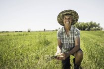 Жінка в полі в сонячному капелюсі тримає спаржу, дивлячись на камеру посміхаючись — стокове фото