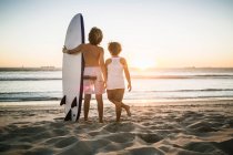 Dos chicos jóvenes de pie en la playa, con tabla de surf, mirando al océano, vista trasera - foto de stock