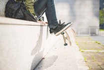 Vita verso il basso vista del giovane skate boarder urbano maschile seduto sul muro flipping skateboard con i piedi — Foto stock