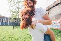 Jovens gêmeos hipster masculinos com barbas vermelhas piggy apoio no parque — Fotografia de Stock