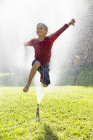Мальчик прыгает через разбрызгиватель в саду — стоковое фото