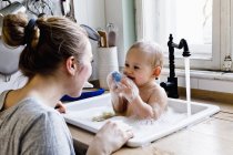 Baby boy sucking sponge whilst bathing in kitchen sink — Stock Photo