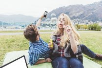 Parejas jóvenes en la manta de picnic soplando y fotografiando burbujas, Lago de Como, Italia - foto de stock