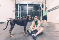 Jovem macho cross trainer com cão amarrando cadarços treinador no ginásio — Fotografia de Stock