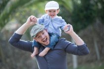 Baby boy wearing baseball cap sitting on father shoulders looking at camera smiling — Stock Photo