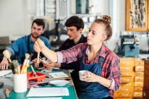 Young craftswoman holding coffee cup and reaching out for paintbrush in creative print studio, with two young craftsmen working in background — Stock Photo