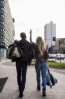 Back view of walking caucasian woman and African ethnicity man in street, couple in love carrying baggage, woman gesturing peace sign with fingers — Stock Photo