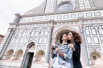 Low angle view of lesbian couple holding digital camera kissing on cheek in front of church, Piazza Santa Maria Novella, Florence, Tuscany, Italy — Stock Photo