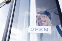 Mujer colocando letrero abierto en la puerta de cristal sonriendo - foto de stock