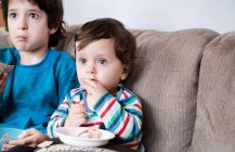 Junge und großer Bruder sitzen auf Sofa und schauen fern, während sie einen Snack essen — Stockfoto
