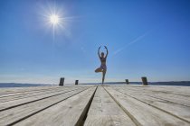 Mid distant view of woman on pier, standing on one leg arms raised — Stock Photo
