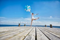 Young woman dancing on wooden pier, holding bunch of balloons — Stock Photo