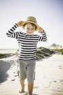 Young boy walking along beach, wearing straw hat, smiling — Stock Photo