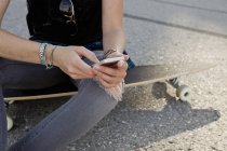 Mid section of female skateboarder sitting on skateboard texting on smartphone — Stock Photo