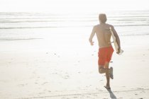 Вид сзади молодого серфера-мужчины, бегущего по освещенному солнцем пляжу, Кейптаун, Западный Кейп, Южная Африка — стоковое фото