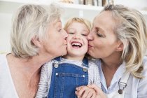 Portrait of girl being kissed on cheek by mother and grandmother in kitchen — Stock Photo