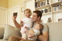 Hombre adulto medio tomando selfie teléfono inteligente con niño y bebé hija en el sofá - foto de stock